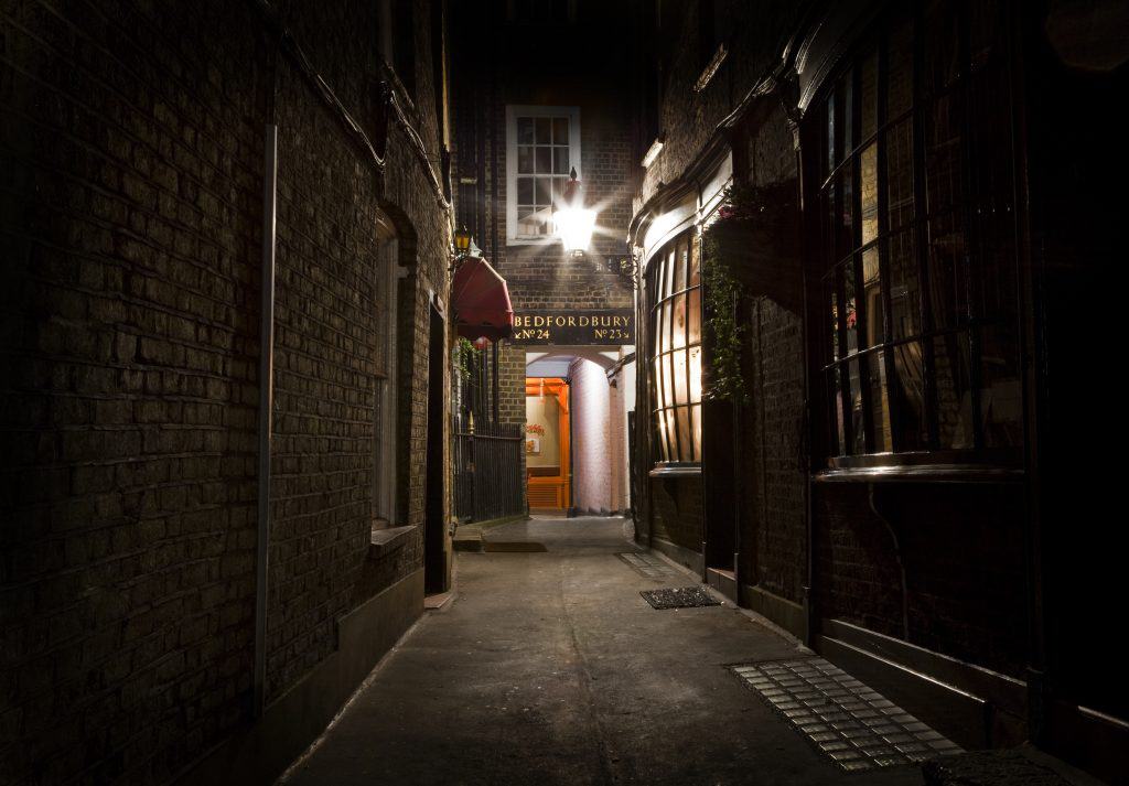 Dimly lit alleyway with a sign reading "Bedfordbury," leading to an illuminated entrance at the end, surrounded by brick walls and narrow passage.