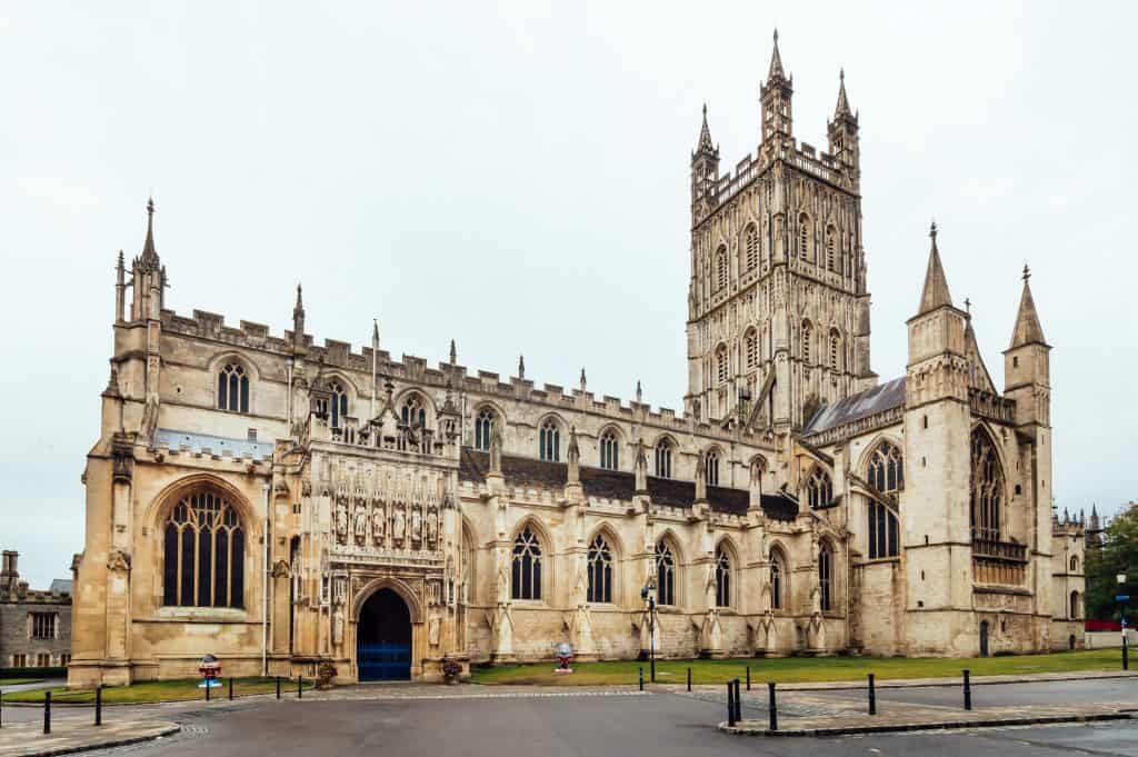 A large Gothic-style stone cathedral with intricate architectural details, featuring a central tower and pointed arches, set against a cloudy sky. Gloucester Cathedral in England.