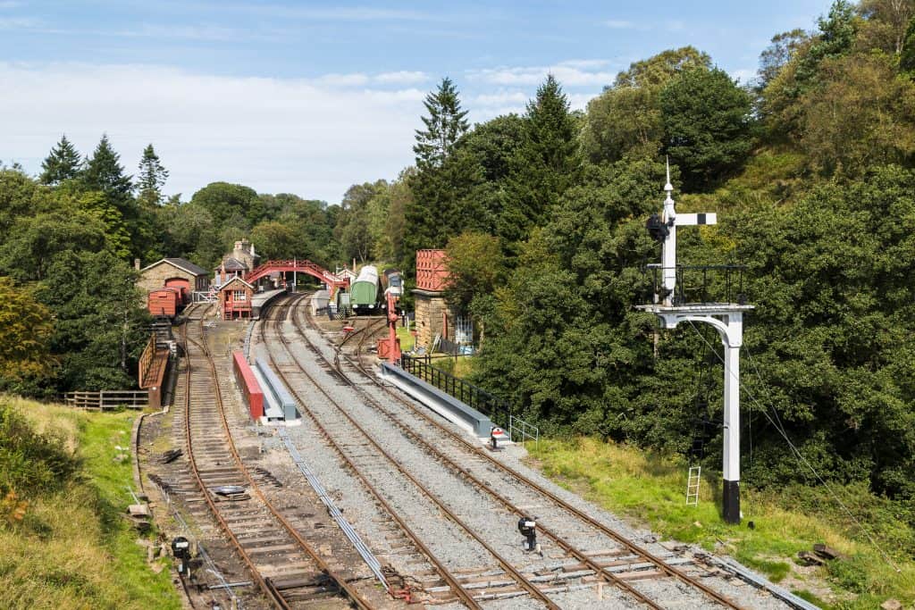 Scenic railway station surrounded by trees, featuring multiple tracks, a signal post, red brick buildings, and a footbridge.
