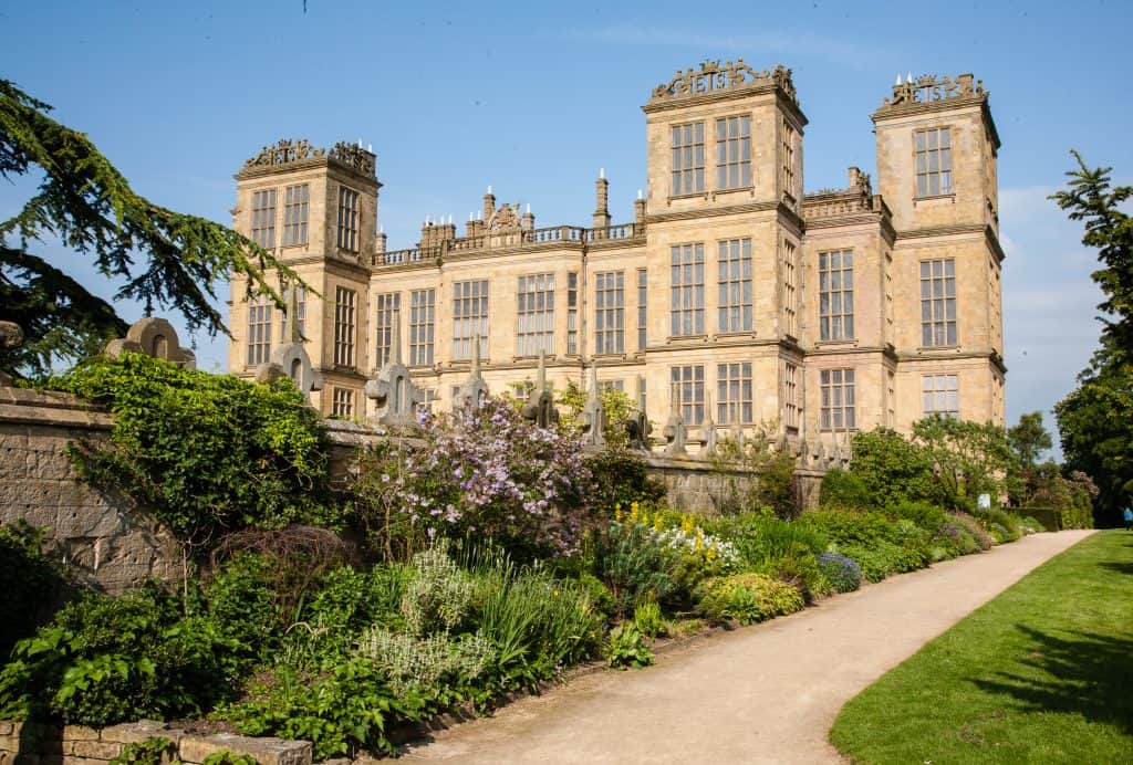 Historic stone mansion with ornate rooftop designs, surrounded by lush gardens and a clear blue sky. Hardwick Hall in England.