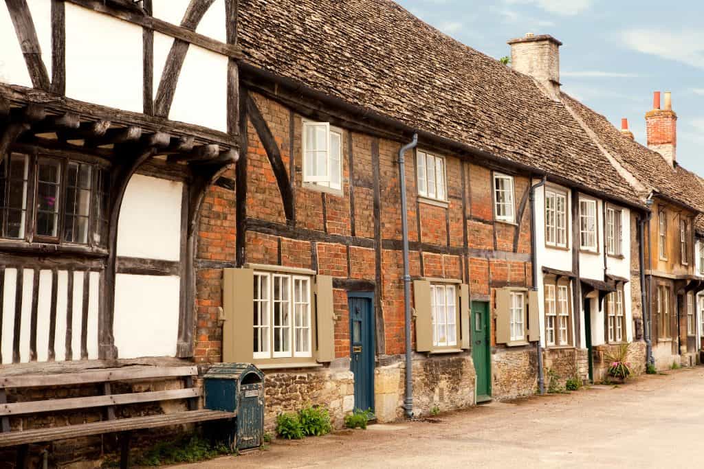 Row of charming, old English cottages with timber frames, brickwork, and shingled roofs under a blue sky.