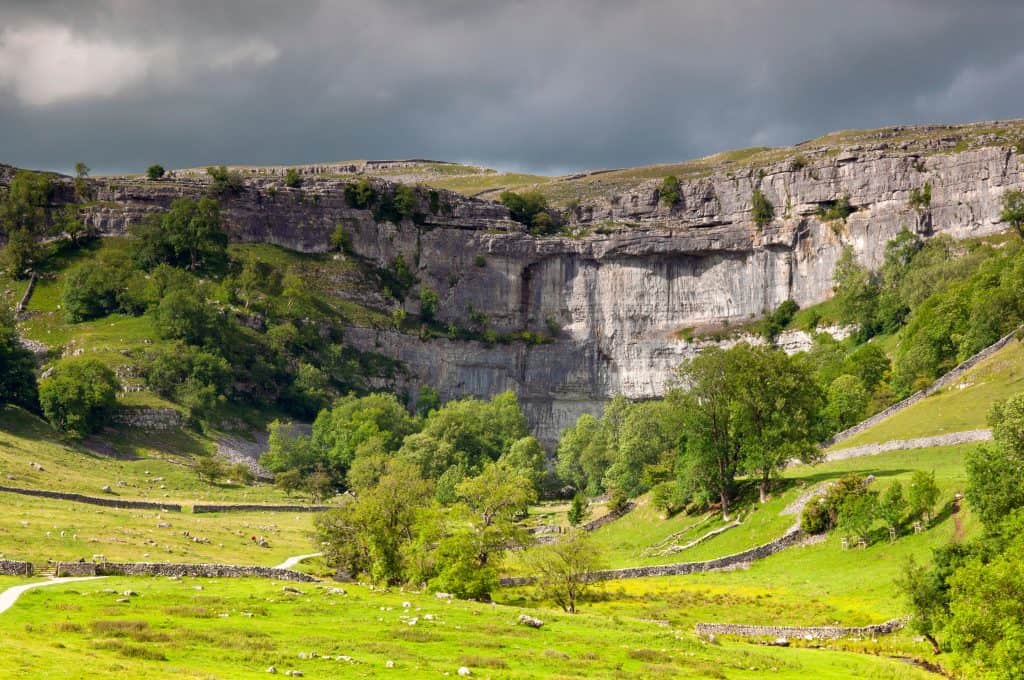 A steep limestone bluff surrounded by green fields and trees under a cloudy sky. Malham Cove in England.
