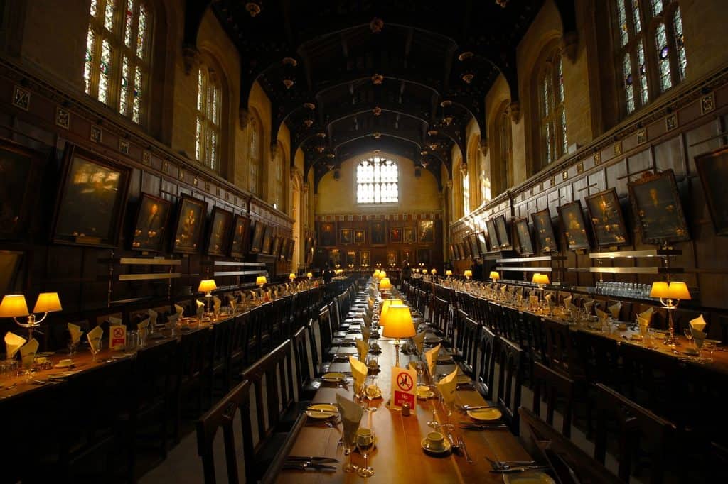 A long, ornate dining hall with high ceilings, wooden paneling, portraits on the walls, and tables set with white napkins and lamps.
