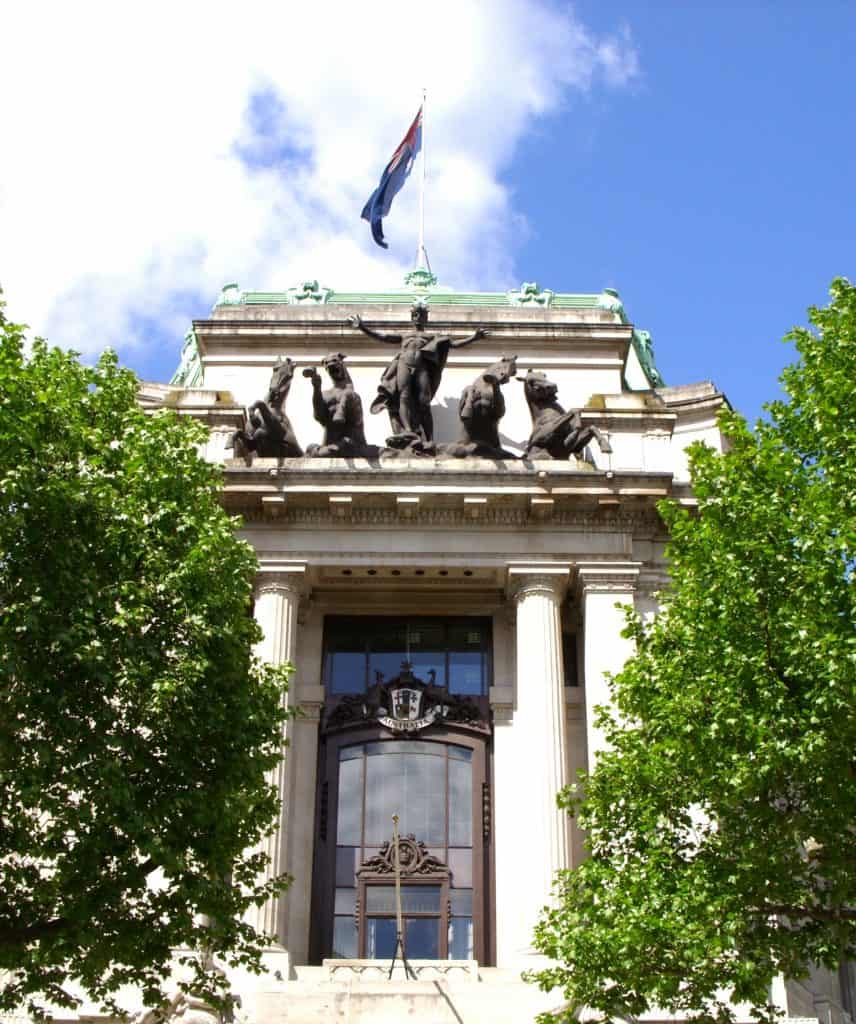 Entrance of a classical building with sculptures above the columns, a flag on top, and green trees on either side.
