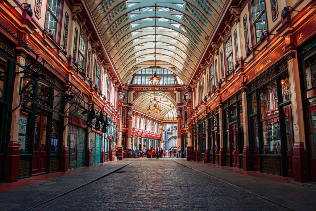 A vibrant, covered market arcade with intricate architecture, arched glass ceiling, and shops on both sides. Warm lighting enhances the colorful facades and cobblestone path.