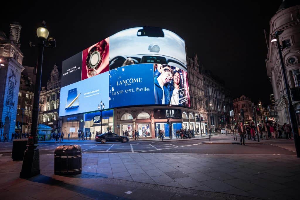 Piccadilly Circus at night with illuminated advertising screens, including a Lancôme ad. Streets are mostly empty, with some pedestrians visible.