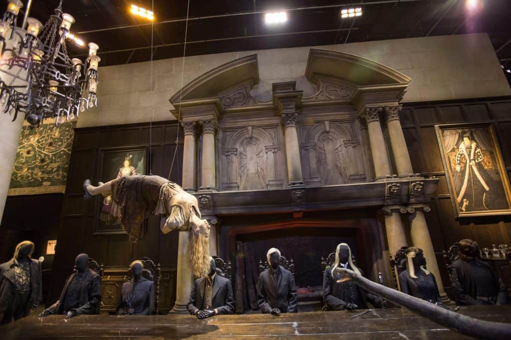 A dimly lit room with a levitating figure above a table, reminiscent of scenes from Harry Potter things to do in London, surrounded by dark-clothed, faceless figures. Ornate dark architectural details and framed artwork adorn the background.