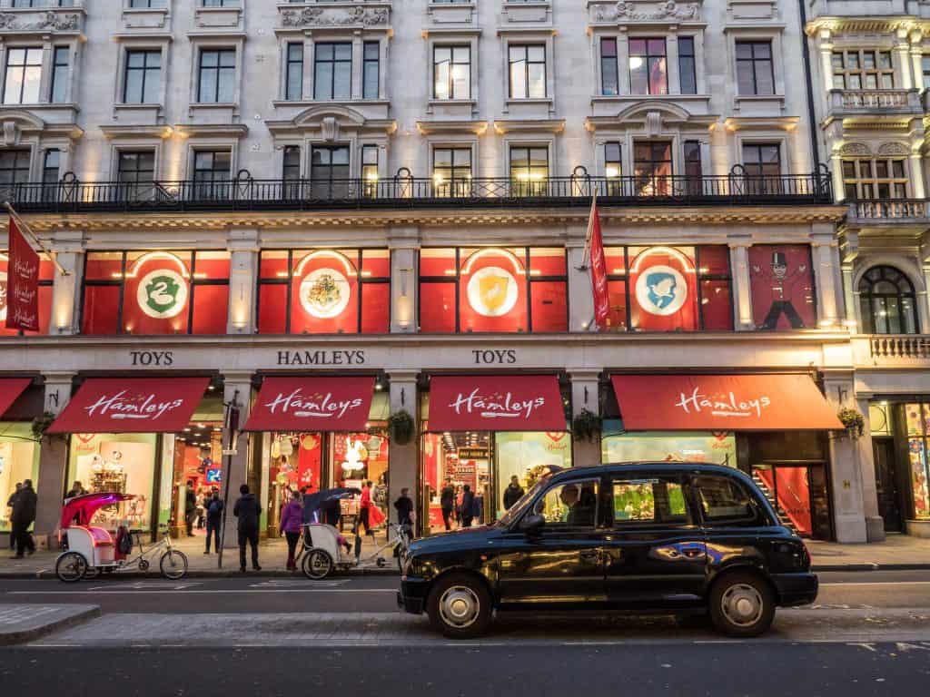 Street view of a Hamleys toy store, its windows adorned with Harry Potter displays enchanting pedestrians. A black taxi and two pedicabs bustle along the road in front of the building, capturing the magical spirit of things to do in London.