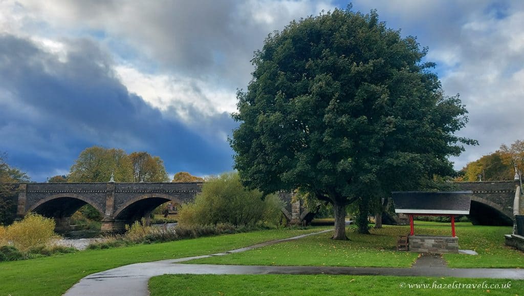 A large tree and a pavilion stand near a stone bridge over a river, with a grassy area and a path in the foreground under a cloudy sky.