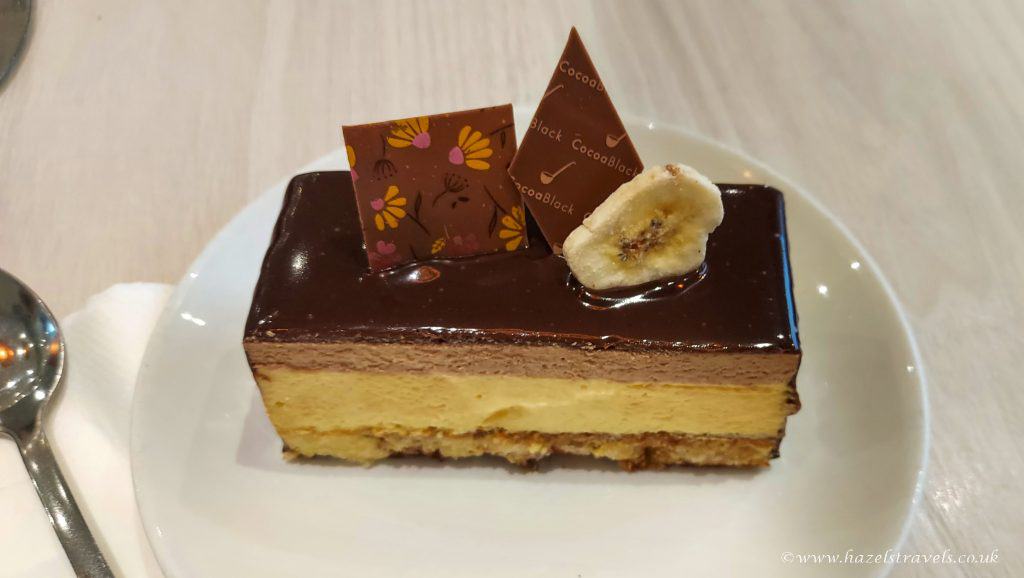 A slice of layered cake with chocolate glaze, decorated with chocolate pieces and a banana slice on top, sits on a white plate beside a spoon.