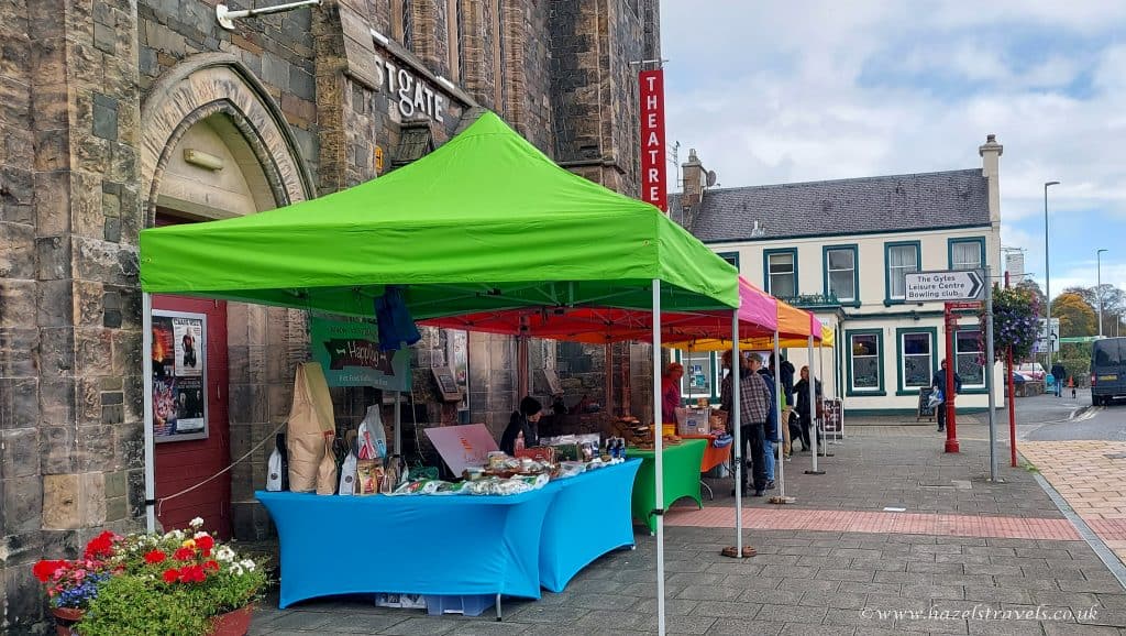 Colorful market stalls with goods for sale are set up along a street near a theater entrance. People are browsing the items.
