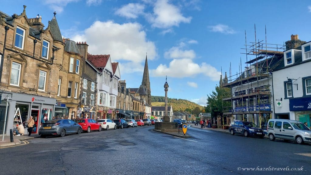 A town street with parked cars, shops, and a pointed church steeple in the background under a partly cloudy sky.