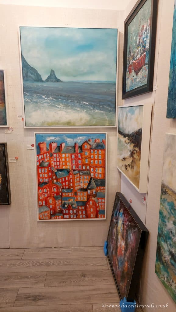 Gallery corner displaying multiple paintings with sea landscapes and abstract cityscapes on a light wooden floor.