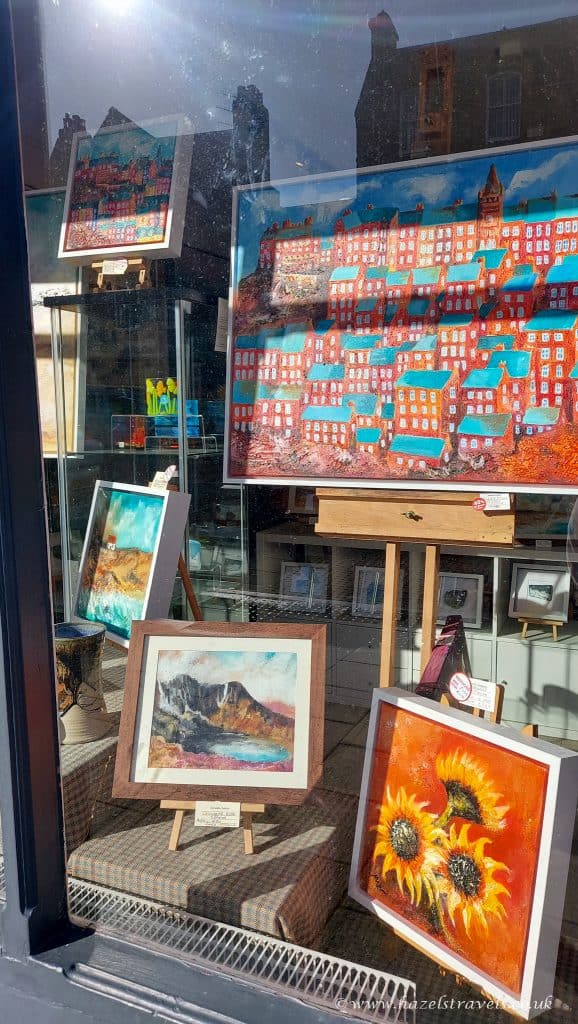 Window display featuring framed paintings, including cityscapes and sunflowers, on easels and shelves. Reflections of nearby buildings seen in the glass.