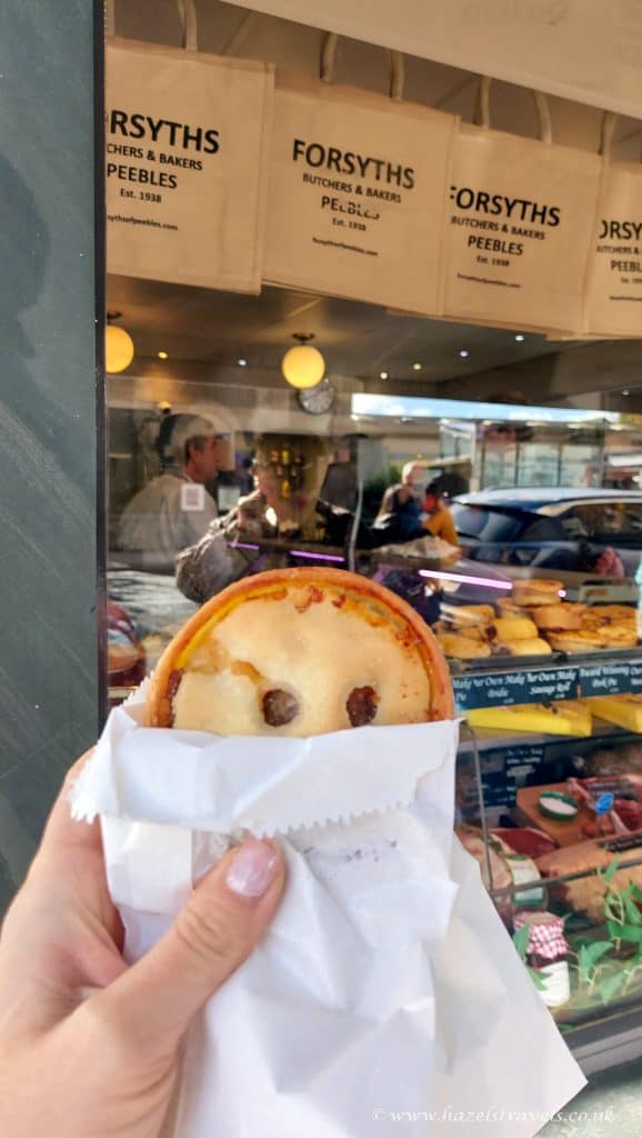 A hand holding a meat pie in front of a bakery window with shopping bags on display and people inside.