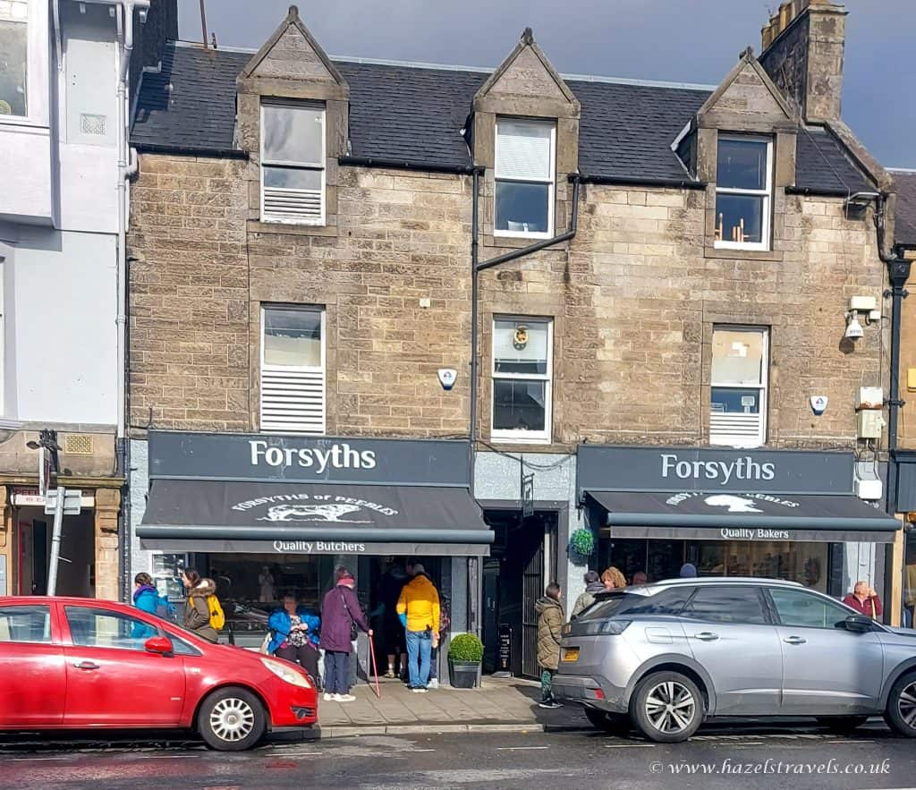 People stand outside Forsyths in Peebles, a building with quality butcher and baker shops on the ground floor. Two cars are parked on the street in front.