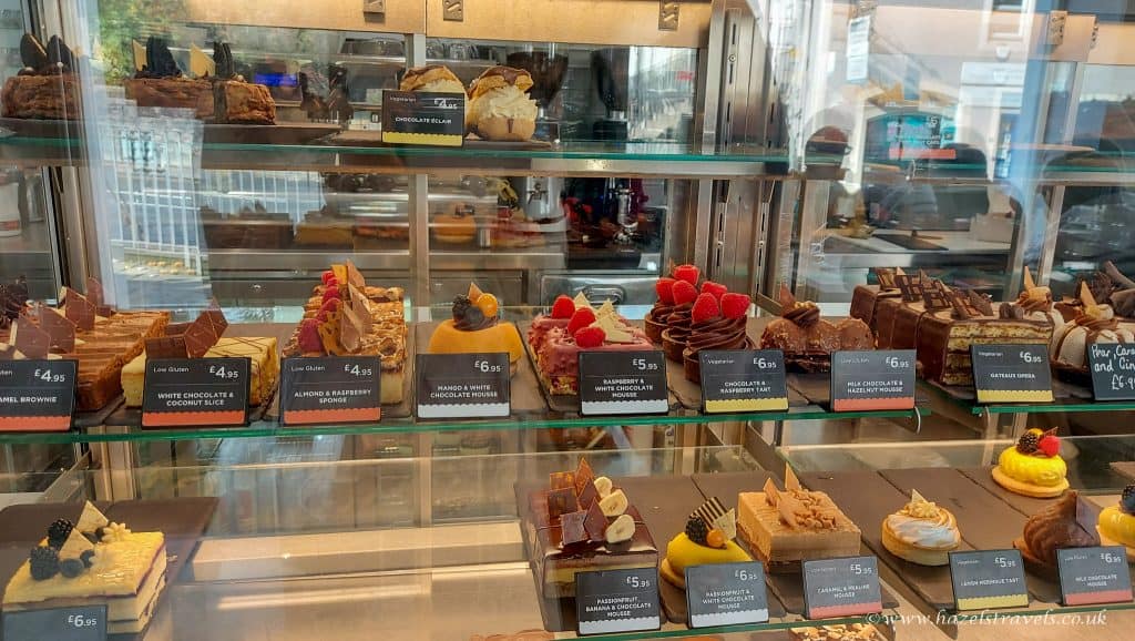 A display case in Peebles is filled with various cakes and pastries, each tagged with a price. Delightful flavors like chocolate, almond, and raspberry are elegantly arranged on the glass shelves.
