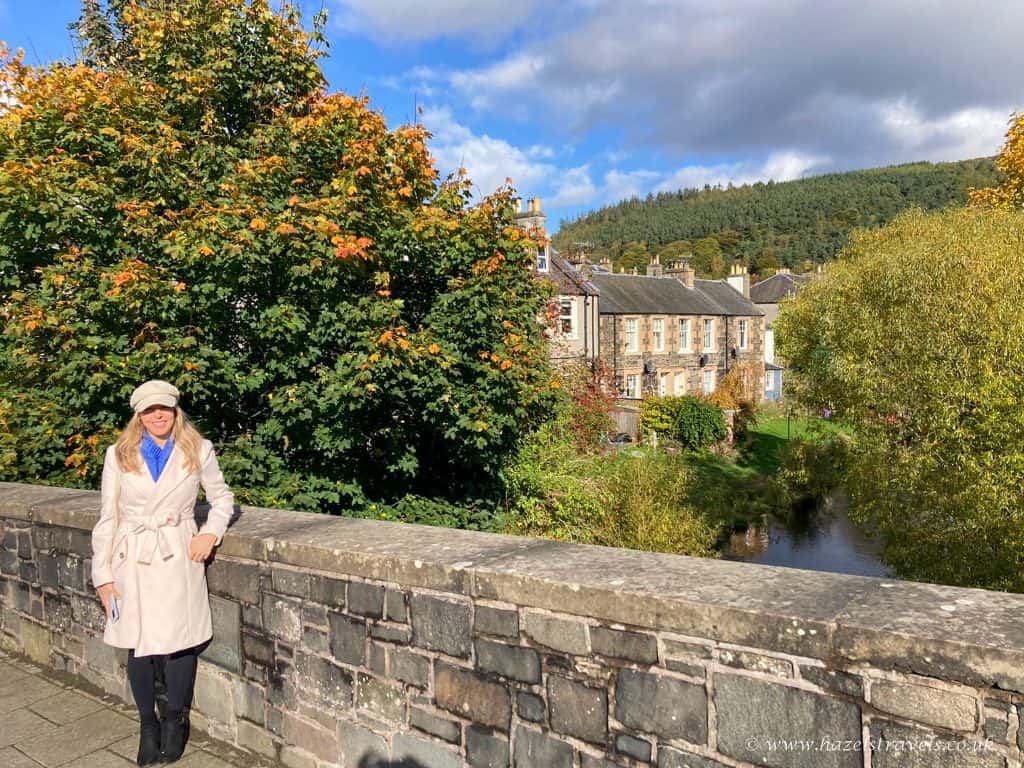 A person in a light coat stands on a stone bridge with trees and buildings in the background. The sky is partly cloudy.