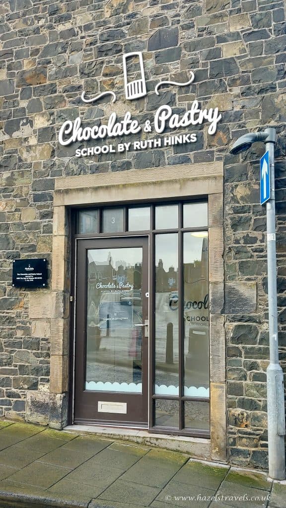 Stone building facade with a glass door. Sign reads "Chocolate & Pastry School by Ruth Hinks" above. A plaque is mounted on the stone wall beside the door.