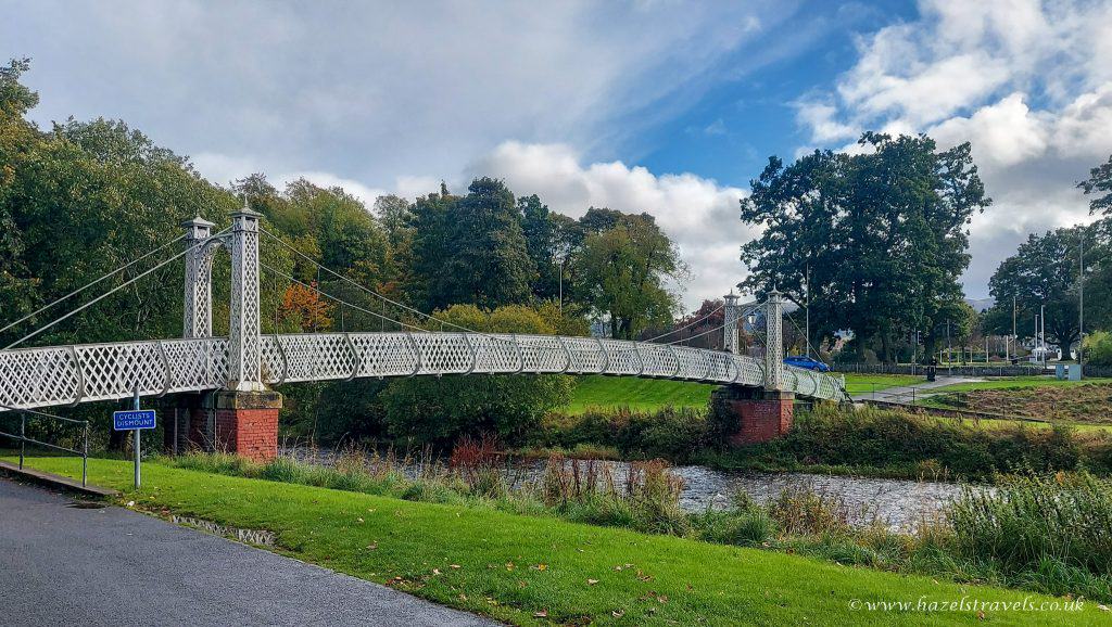 A white suspension footbridge crosses a river, surrounded by green grass and trees under a cloudy sky. A small sign reads "Dumfries.