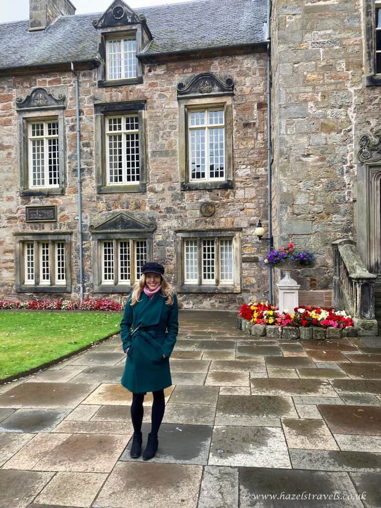 A person in a green coat stands smiling in front of a historic stone building with tall windows, surrounded by a garden with colorful flowers.
