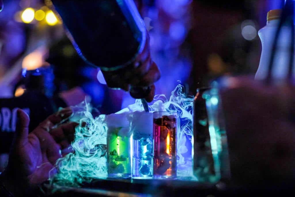 In a dimly lit setting, reminiscent of Harry Potter's world, a hand pours liquid into glowing, steaming test tube glasses.