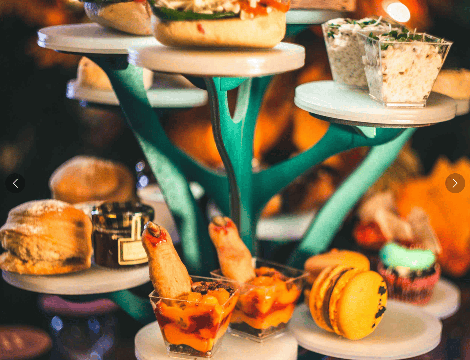 Assortment of food on tiered trays, including sandwiches, dips, scones, macarons, and appetizers in small cups, displayed elegantly.