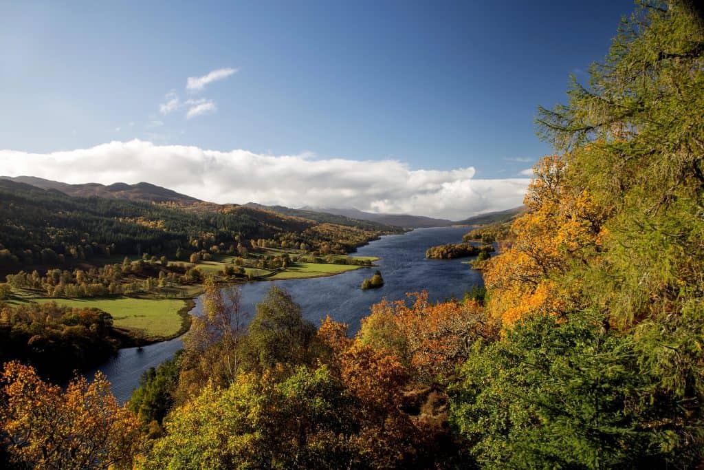 A scenic view of a river winding through a valley in Pitlochry surrounded by autumn trees, under a clear blue sky.