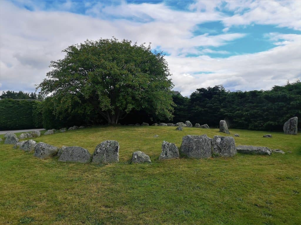 A circular arrangement of standing stones surrounds a tree in a grassy field under a partly cloudy sky.