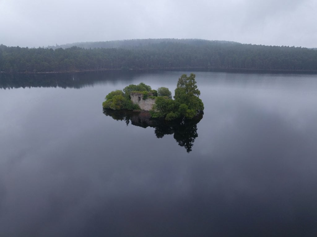 A small, tree-covered island with a stone building is surrounded by a calm, reflective lake, under an overcast sky.