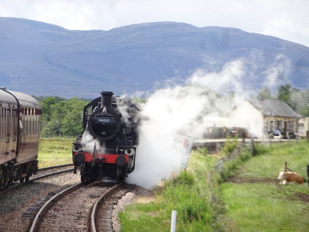 A steam locomotive travels on a curved railway track in a rural landscape with mountains in the background and a building nearby.