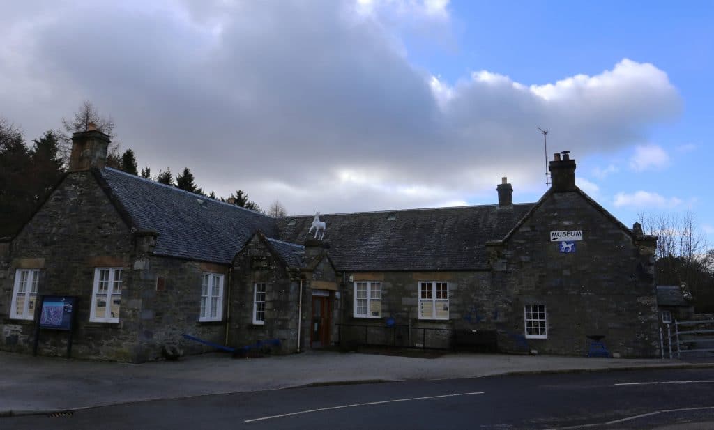 A small gray stone building with a sloped roof, a white cow statue on top, and signs on the front. Surrounded by trees under a cloudy sky.