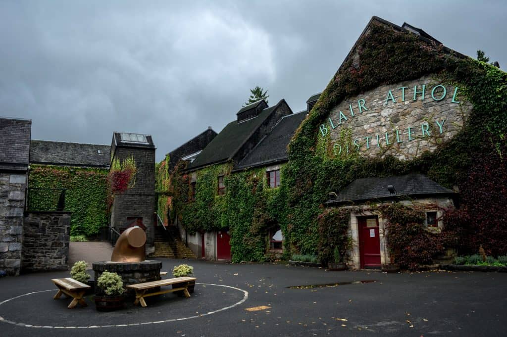 Blair Athol Distillery with ivy-covered stone buildings, a picnic table, and a copper distillation pot on a cloudy day.