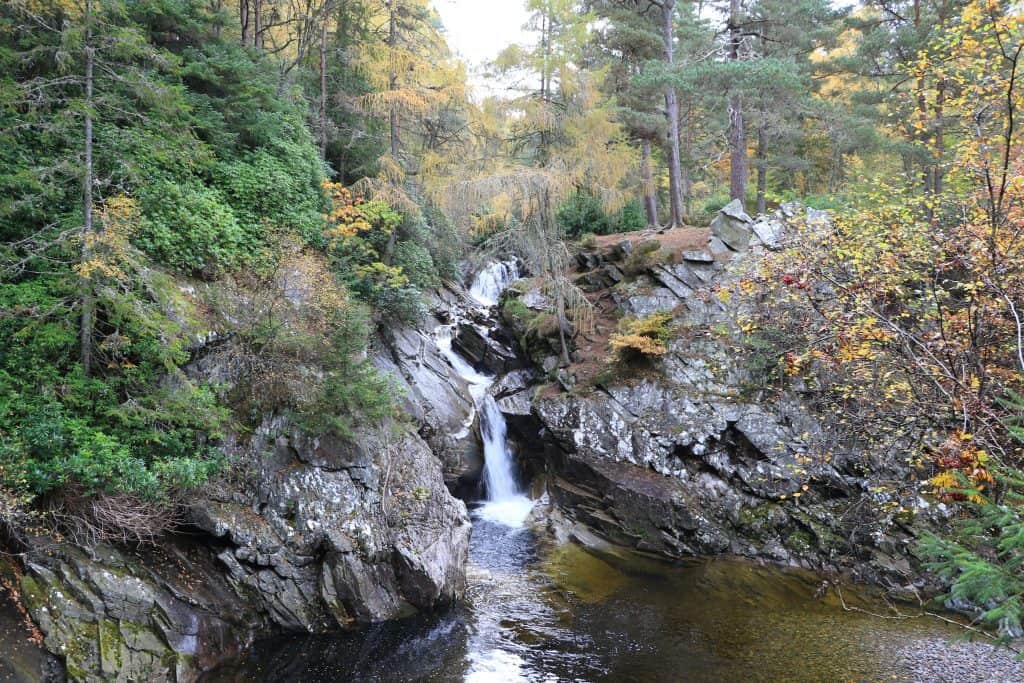 A small waterfall flows between rocky cliffs surrounded by trees with autumn foliage.