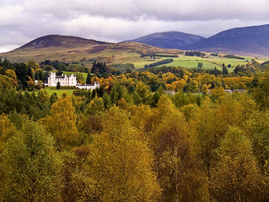A white castle surrounded by dense green and yellow trees, with rolling hills and mountains in the background under a cloudy sky.