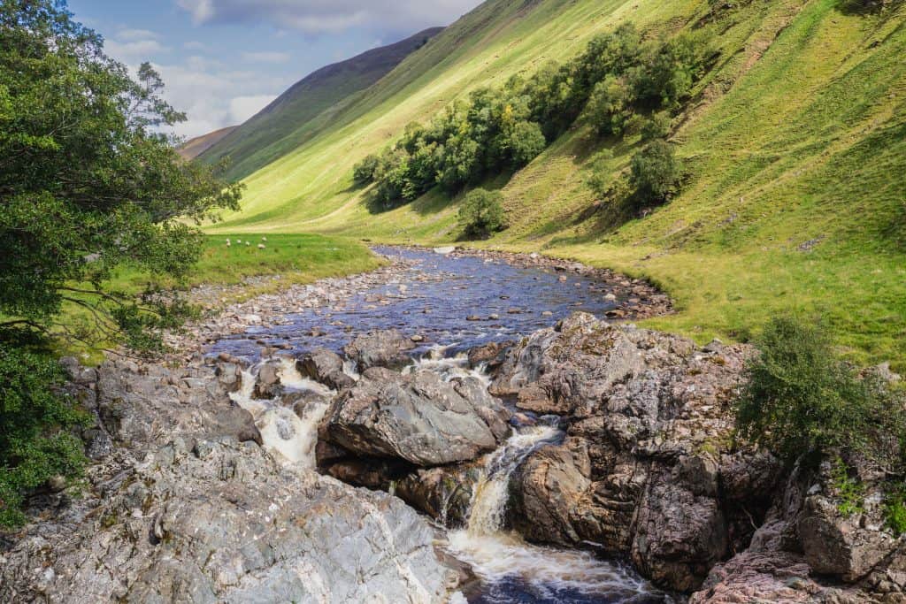 A small stream flows over rocks in a green valley with grassy hills and trees under a partly cloudy sky in Glen Tilt near Blair Atholl.