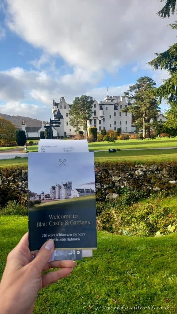 A hand holds a brochure in front of Blair Castle & Gardens, with an expansive lawn and pine trees under a partly cloudy sky.