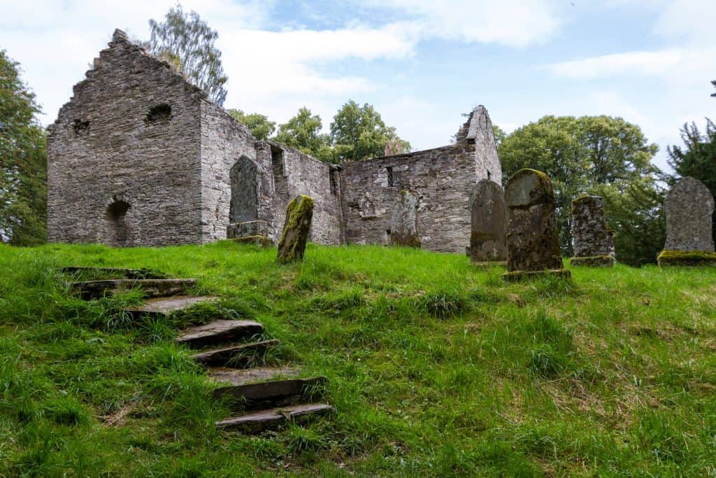 Ruins of an old stone building surrounded by gravestones on a grassy hillside, with trees in the background inthe grounds of Blair Atholl Castle.