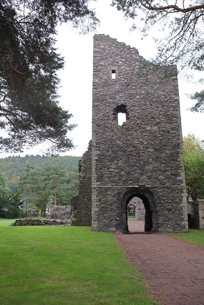 A tall, weathered stone tower in Peebles, Scotland with a large archway stands on a grassy field, surrounded by trees.