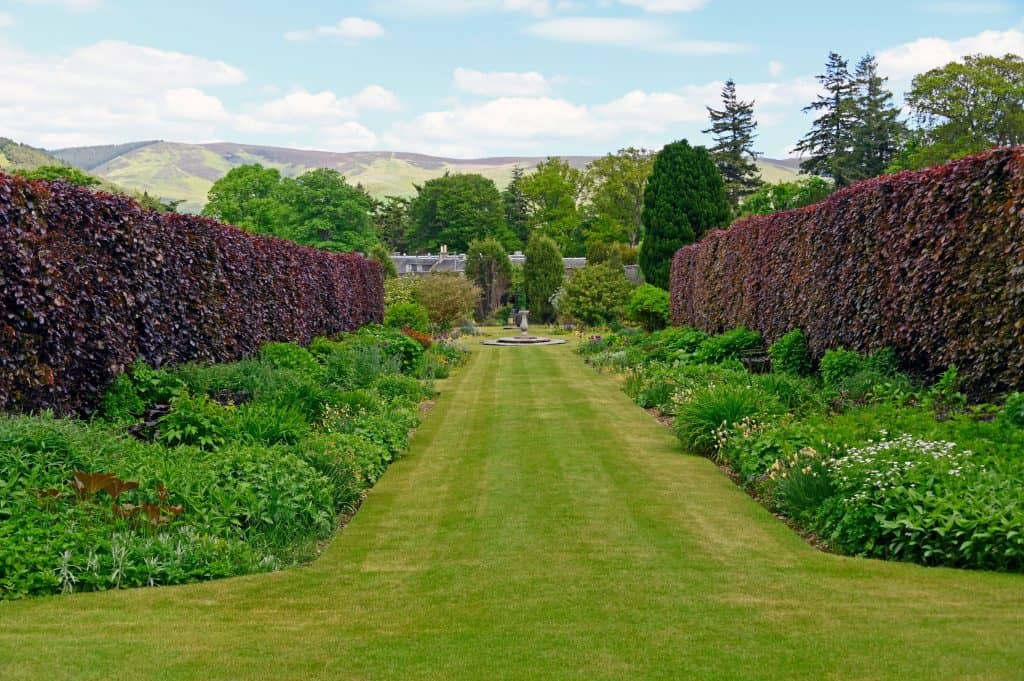 A neatly trimmed grass path runs between two tall hedges, surrounded by lush green gardens, trees, and hills in the background.