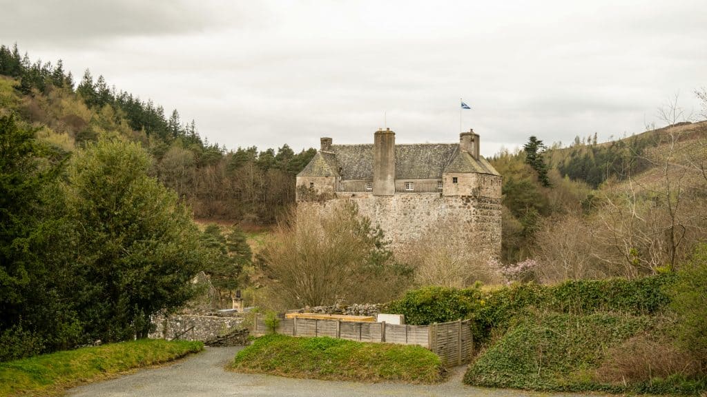 The historic Neidpath Castle with a flag atop, surrounded by trees, sits under a cloudy sky. A gravel path leads to the entrance.