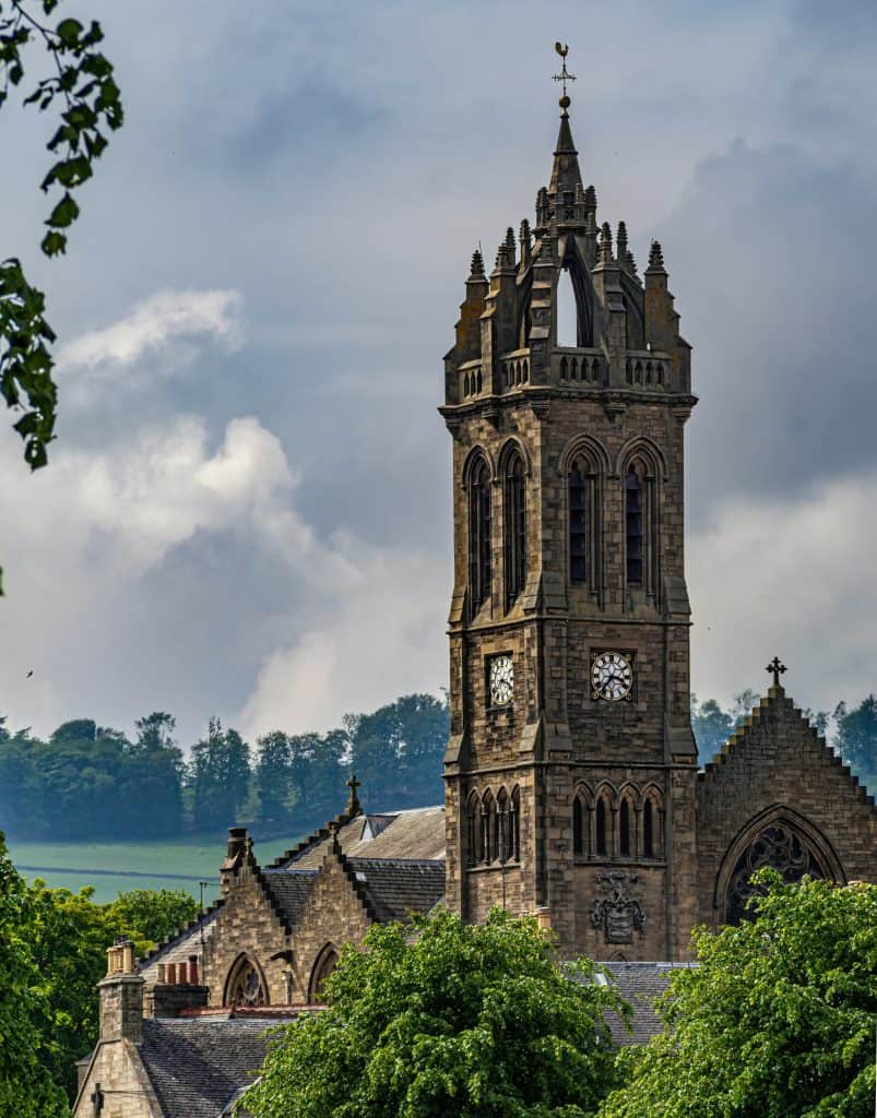 Peebles Old Parish Church with a tall clock tower surrounded by green trees under a cloudy sky.