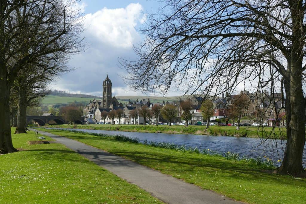 A riverside path with trees, leading to a town with a prominent church tower in Peebles, Scotland. Green fields and cloudy sky in the background.
