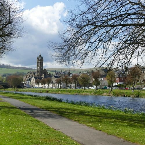 A riverside path with trees, leading to a town with a prominent church tower in Peebles, Scotland. Green fields and cloudy sky in the background.
