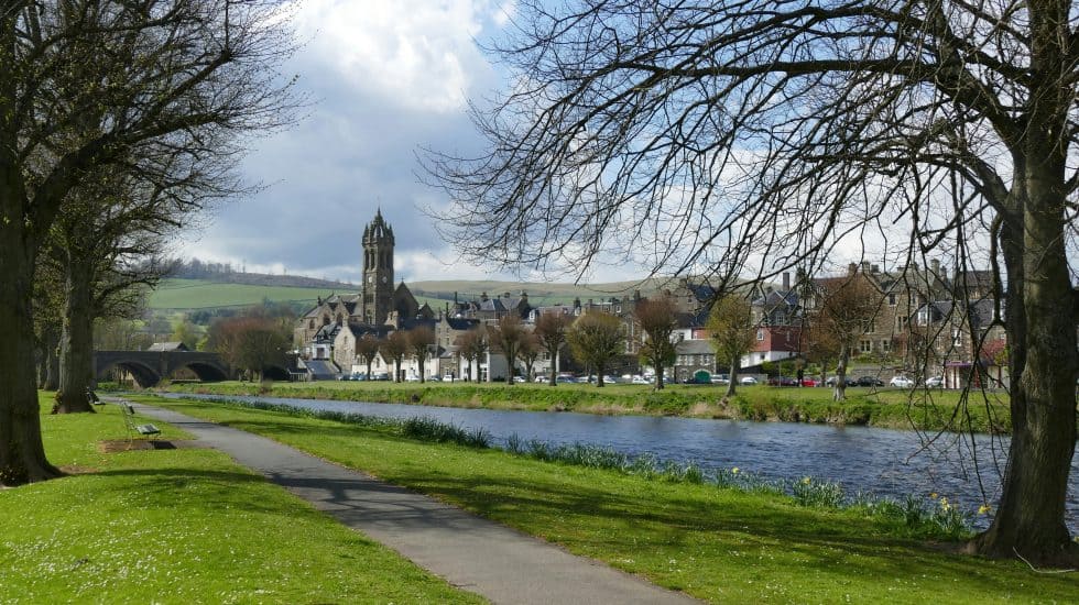 A riverside path with trees, leading to a town with a prominent church tower in Peebles, Scotland. Green fields and cloudy sky in the background.