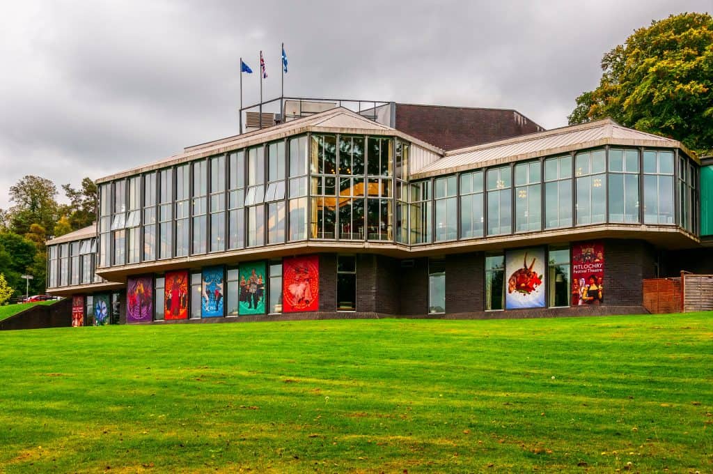 A modern glass and brick building in Pitlochry with colorful banners on the lower level, surrounded by a grassy lawn and trees. Flags are displayed on the roof against a cloudy sky.