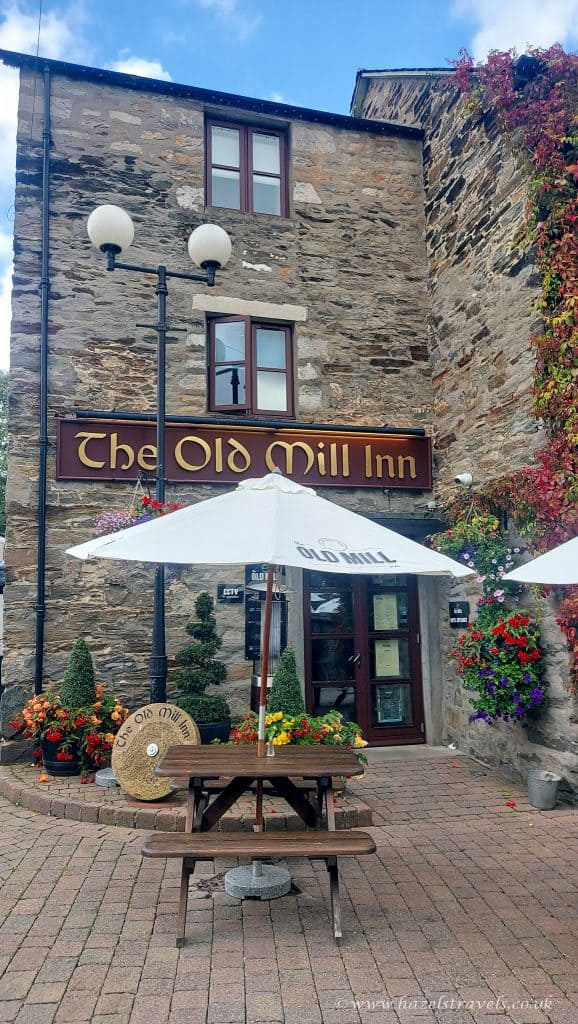 Stone building of The Old Mill Inn with two windows above the entrance, surrounded by colorful flowers and outdoor seating with umbrellas.