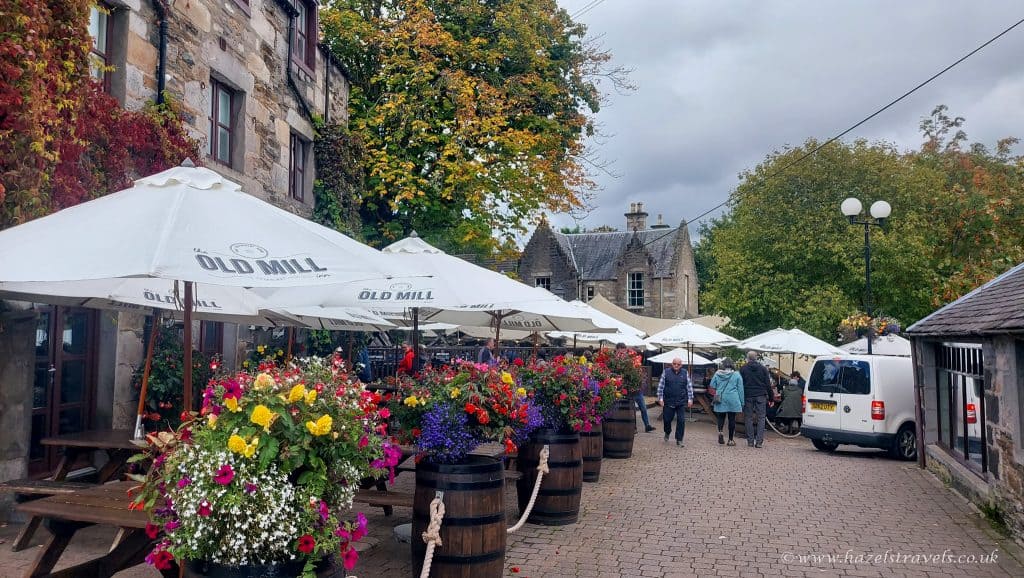 Outdoor seating area with white umbrellas labeled "Old Mill," surrounded by colorful flower barrels. People walk along a cobblestone path near stone buildings and leafy trees.
