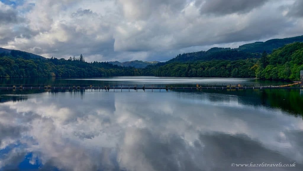 A serene lake reflects clouds and a forested hill, with a narrow bridge crossing the water under a cloudy sky.