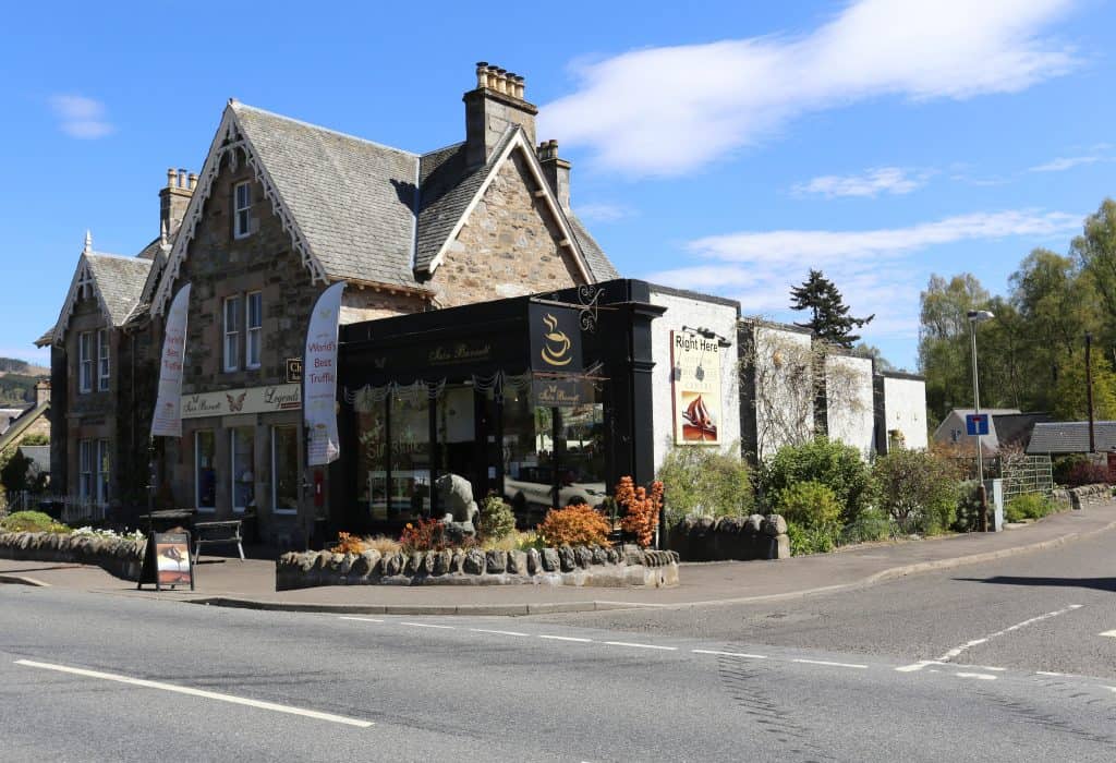 Street view of a stone building housing a restaurant with large windows, decorative signs, and colorful plants in front. A road and clear sky are visible.